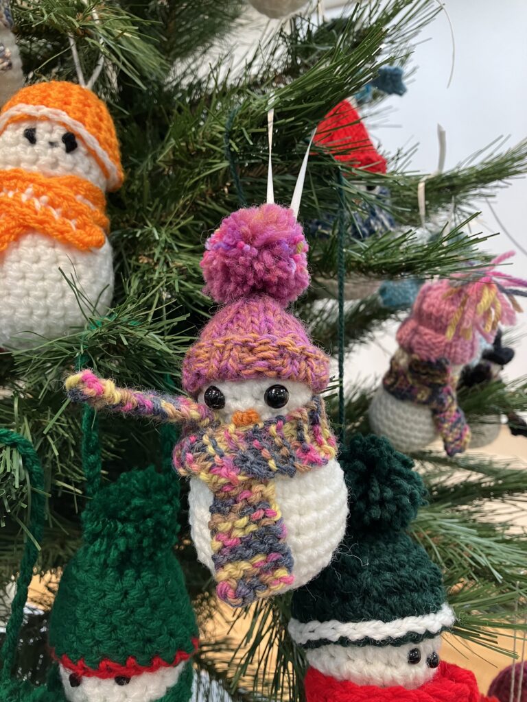 A closeup image of one crochet snowman hanging on a tree, round and fluffy, this one has a pink pom hat on with a patterned pink, grey, and yellow scarf. Behind this snowman is another with an orange pom hat and orange and yellow scarf.