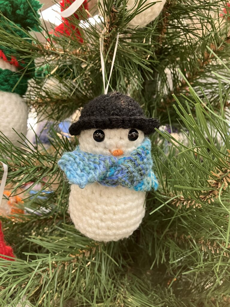 A closeup image of one crochet snowman hanging on a tree, round and fluffy, this one has a black boiler hat on with a patterned blue and purple scarf.