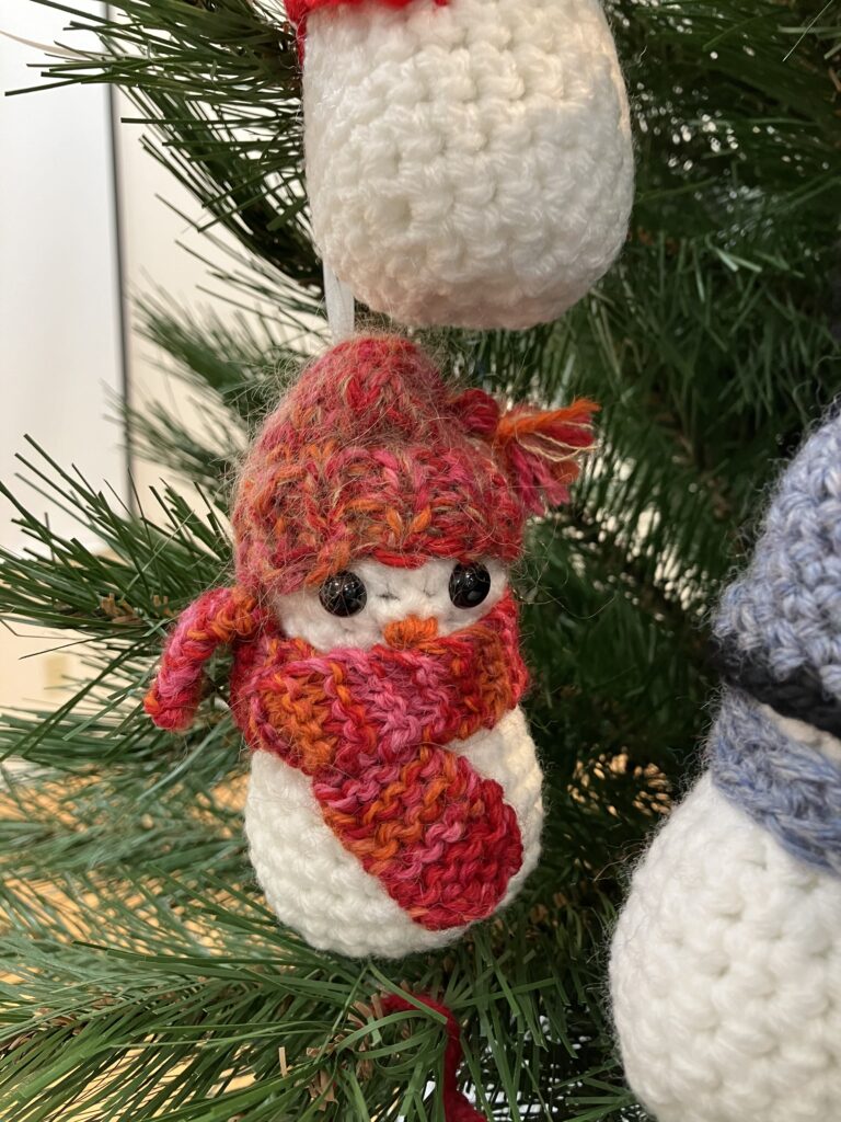 A closeup image of one crochet snowman hanging on a tree, round and fluffy, this one has a red pom hat on with a patterned red scarf.