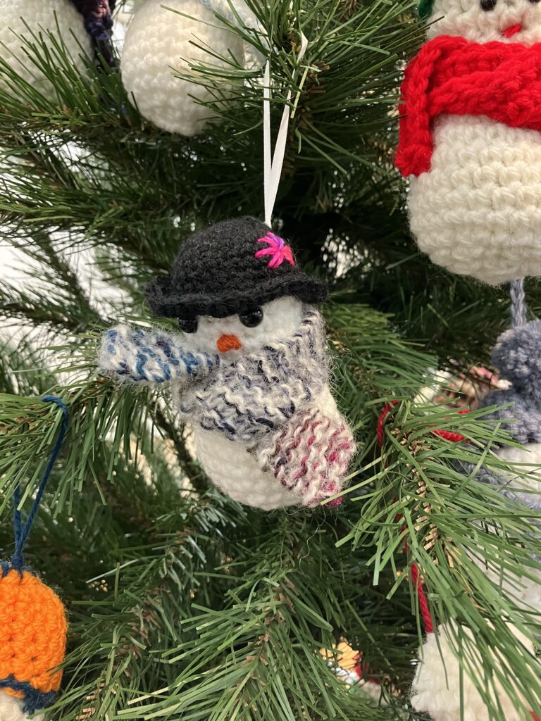 A closeup image of one crochet snowman hanging on a tree, round and fluffy, this one has a black boiler hat with a purple flower on with a patterned grey and red scarf. This snowman is giving the camera a sassy look