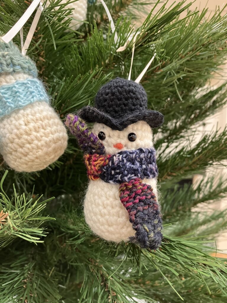 A closeup image of one crochet snowman hanging on a tree, round and fluffy, this one has a tall black fancy boiler hat that curves in a V shape on the front of the cap on with a patterned grey, burnt orange, and red scarf. This snowman is looks like a detective out for a day's work.