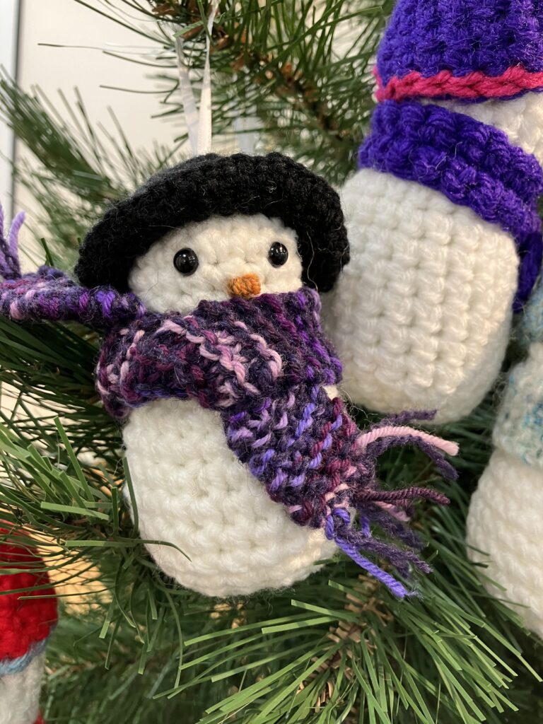 A closeup image of one crochet snowman hanging on a tree, round and fluffy, this one has a black pom hat on with a patterned purple scarf.